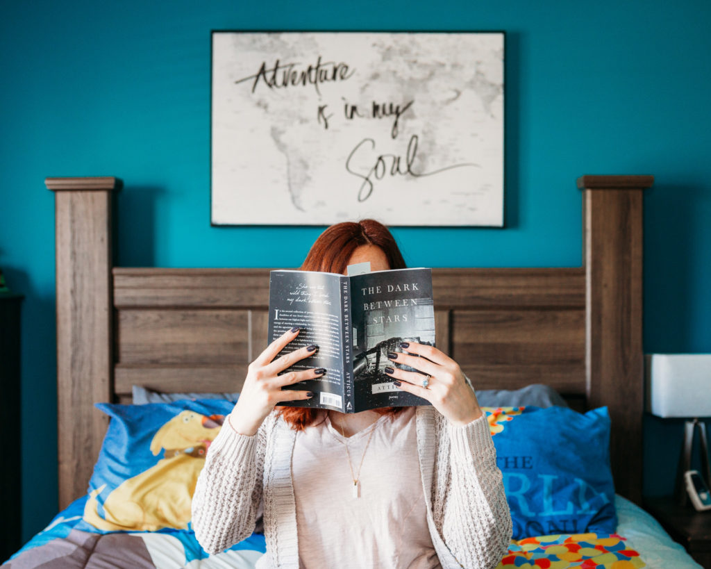 Red headed woman sitting cross legged on bed holding a book in front of her face